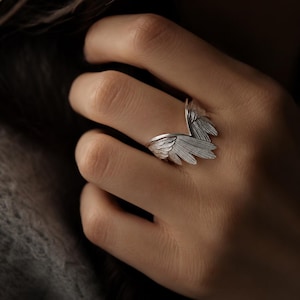 Peut inclure: Une bague en argent avec un motif d'aile. La bague présente des textures de plumes détaillées, avec une aile s'étendant vers le haut et l'autre enroulée autour du doigt. La bague est portée sur une main, mettant en valeur le design et le savoir-faire du bijou.