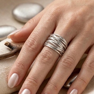 Peut inclure: Une bague en argent avec un design tressé est présentée sur un doigt. La bague est composée de plusieurs brins d'argent, créant un aspect texturé et superposé. La main est visible, avec des ongles polis. Le fond comprend de petites pierres argentées.