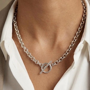 Peut inclure: Collier argenté avec une chaîne épaisse et un fermoir circulaire avec une barre. Le collier est porté sur une chemise blanche à col et un blazer beige, créant une esthétique minimaliste. Le bijou est une pièce forte.