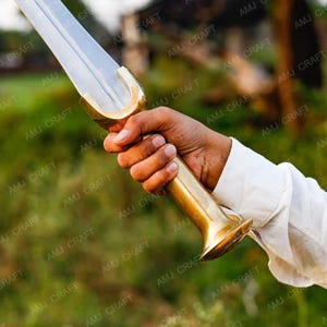 Op de afbeelding: Een zwaard met een gepolijst zilveren lemmet en een goudkleurige handgreep. De handgreep heeft een gebogen ontwerp en wordt in een hand gehouden, met een witte mouw zichtbaar. Het zwaard is een traditioneel wapen.