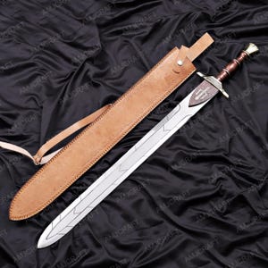 May include: A long, silver sword with a brown handle and a leather sheath. The sword has a decorative hilt and a blade with etched designs. The sheath is tan and has a strap. The sword is displayed on a black, textured surface.