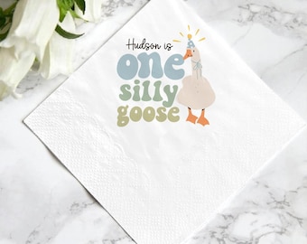 May include: White square paper napkin with the text "Hudson is one silly goose" in blue and green. A cartoon goose wearing a party hat is printed on the napkin. The napkin is on a white and gray marble surface.