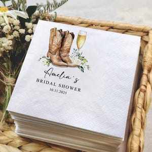 May include: White paper napkins with a diamond pattern, featuring a watercolor illustration of brown cowboy boots, a champagne flute, and floral accents. Text reads "Amelia's Bridal Shower 10.11.2025".