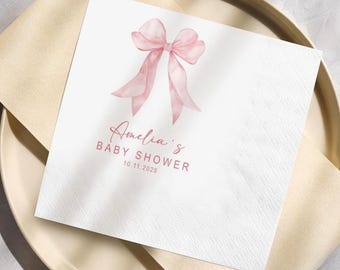 May include: White paper napkin with a watercolor-style pink bow and the text "Amelia's BABY SHOWER 10.11.2025". The napkin is placed on a beige plate and a beige napkin.