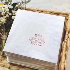May include: White paper napkins with a red swan design and the text "20.11.25" and "Olivia + David". The napkins are stacked in a woven basket, suitable for a wedding or special event.
