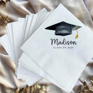 Peut inclure: Serviettes de graduation blanches avec un mortier noir et une illustration de gland dor&eacute;. Le nom "Madison" et "Classe de 2025" sont imprim&eacute;s en script noir. Une pile de serviettes pli&eacute;es est visible.