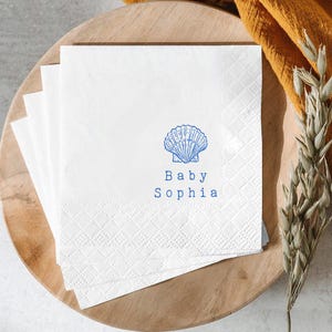 Może przedstawiać: Stos białych papierowych serwetek z niebieską ilustracją liniową muszli umieszczoną nad niebieskim napisem Baby Sophia. Serwetki mają teksturowaną krawędź z wzorem w romby oraz gładki środek.
