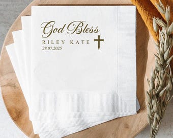 May include: White paper napkins with the words "God Bless", the names "Riley Kate", and the date "28.07.2025" printed in a gold color. A gold cross is also printed on the napkins. The napkins are stacked on a wooden plate.