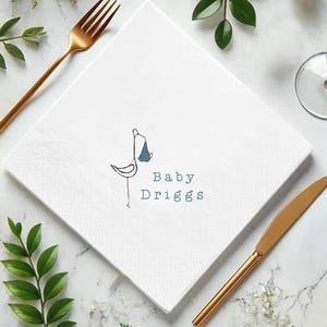 May include: White square paper napkin with a stork carrying a baby in a blue bundle. The text "Baby Driggs" is printed in blue. A gold fork and knife are on the sides, with greenery.