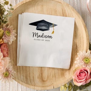 Peut inclure: Serviettes de graduation blanches avec une illustration &agrave; l'aquarelle d'un chapeau de remise de dipl&ocirc;me et d'un gland. Le nom "Madison" et "Classe de 2025" sont imprim&eacute;s en script noir. Serviettes sur un plateau en bois avec des d&eacute;corations florales.