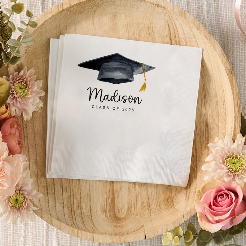 Peut inclure: Serviettes de graduation blanches avec une illustration &agrave; l'aquarelle d'un chapeau de remise de dipl&ocirc;me et d'un gland. Le nom "Madison" et "Classe de 2025" sont imprim&eacute;s en script noir. Serviettes sur un plateau en bois avec des d&eacute;corations florales.