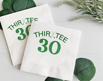 May include: Two white paper napkins with the text "THIRTEE 30" in green, with a golf club graphic. The napkins are surrounded by green leaves and sprigs, suggesting a golf-themed party or event.