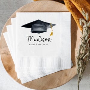 Peut inclure: Serviettes de graduation blanches avec une illustration &agrave; l'aquarelle d'un chapeau de remise de dipl&ocirc;me noir et d'un gland dor&eacute;. Le nom "Madison" et "Classe de 2025" sont imprim&eacute;s en script noir. Serviettes empil&eacute;es.