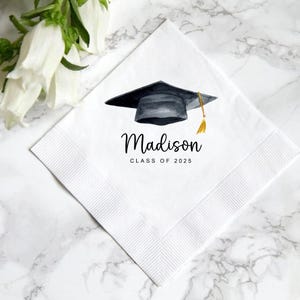 Peut inclure: Serviette carr&eacute;e blanche avec un motif de toque de graduation &agrave; l'aquarelle. La toque est noire avec un gland dor&eacute;. Le nom "Madison" et "CLASS OF 2025" sont imprim&eacute;s en script noir. La serviette a une bordure textur&eacute;e.