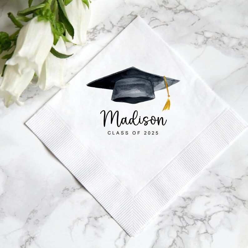 Peut inclure: Serviette carr&eacute;e blanche avec un motif de toque de graduation &agrave; l'aquarelle. La toque est noire avec un gland dor&eacute;. Le nom "Madison" et "CLASS OF 2025" sont imprim&eacute;s en script noir. La serviette a une bordure textur&eacute;e.