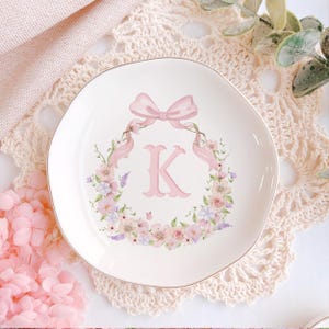 May include: A white ceramic plate with a gold rim, featuring a pink floral wreath and a pink letter "K". A pink bow and ribbons are at the top. The plate is on a cream-colored lace doily.