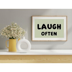 May include: A framed print with a light green background and black text that reads "LAUGH OFTEN". The print is sitting on a white shelf with a yellow vase and a white vase with a ring design.