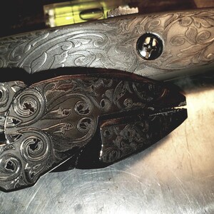 May include: Close-up of a pair of metal pliers and a metal tool, both with intricate floral and scrollwork engravings. The tools are silver-toned and rest on a metallic surface, showcasing detailed craftsmanship.