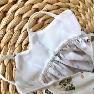 May include: White cotton face mask with a floral print. The mask has elastic ear loops and a white cotton lining.