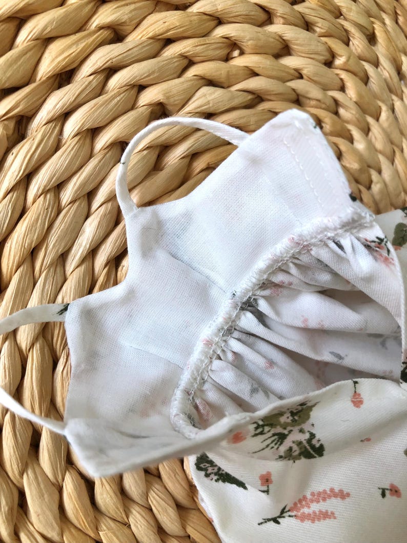 May include: White cotton face mask with a floral print. The mask has elastic ear loops and a white cotton lining.