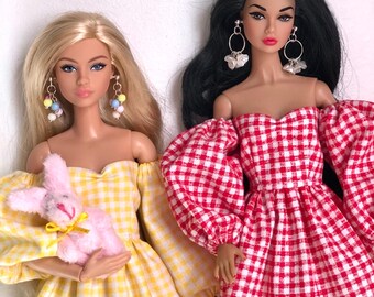 May include: Two fashion dolls with articulated bodies. The doll on the left has blonde hair, wears a yellow gingham off-the-shoulder dress, and holds a small pink plush rabbit. The doll on the right has black hair and wears a red gingham dress.