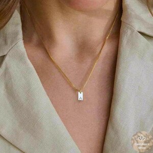 Op de afbeelding: Een gouden ketting met een klein rechthoekig hangertje met de letter 'A' in wit.