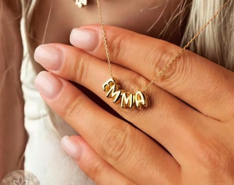 May include: A gold necklace featuring the name "EMMA" spelled out in individual letter beads. The necklace is held by a person with manicured nails. Another necklace is visible in the background.