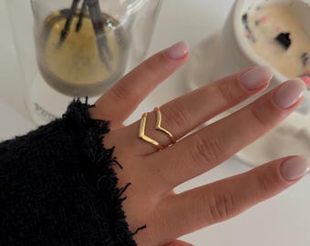May include: A close-up of a hand wearing two gold rings. The rings are stacked on the middle finger and are shaped like a V. The hand is resting on a white surface.