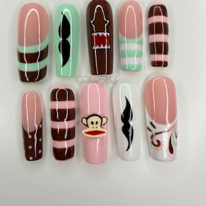 Könnte beinhalten: Ein Set aus zehn künstlichen Nägeln mit verschiedenen Designs. Die Nägel zeigen Farben wie Rosa, Braun, Weiß und Mintgrün. Designs umfassen Streifen, einen Schnurrbart, einen Affen und abstrakte Formen. Der Markenname "Teetree Nails" ist sichtbar.