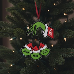 May include: A Christmas tree ornament featuring a green hand holding three Grinch-themed heads with names and Santa hats. The ornament includes a red tag with the year 2025. The ornament hangs from a red ribbon against a backdrop of a Christmas tree.