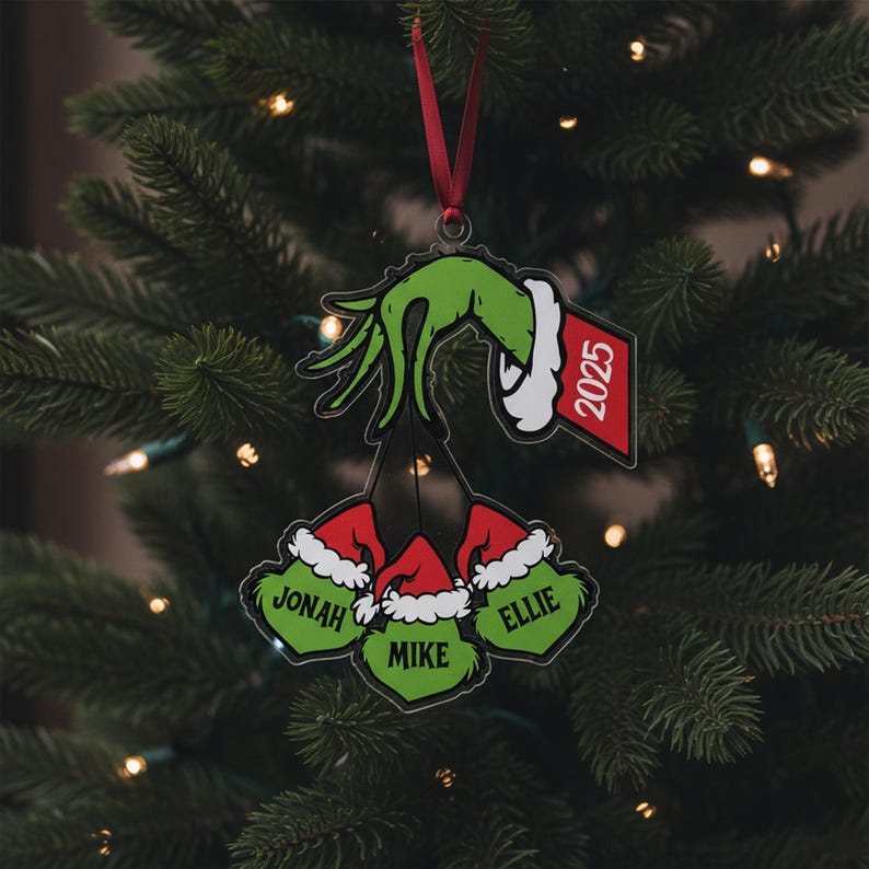 May include: A Christmas tree ornament featuring a green hand holding three Grinch-themed heads with names and Santa hats. The ornament includes a red tag with the year 2025. The ornament hangs from a red ribbon against a backdrop of a Christmas tree.