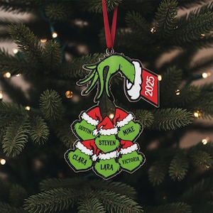 May include: A Christmas tree ornament featuring a green hand with a white cuff holding a cluster of red and white Santa hats. Each hat has a name on it. A red tag with the year 2025 hangs from the hand.