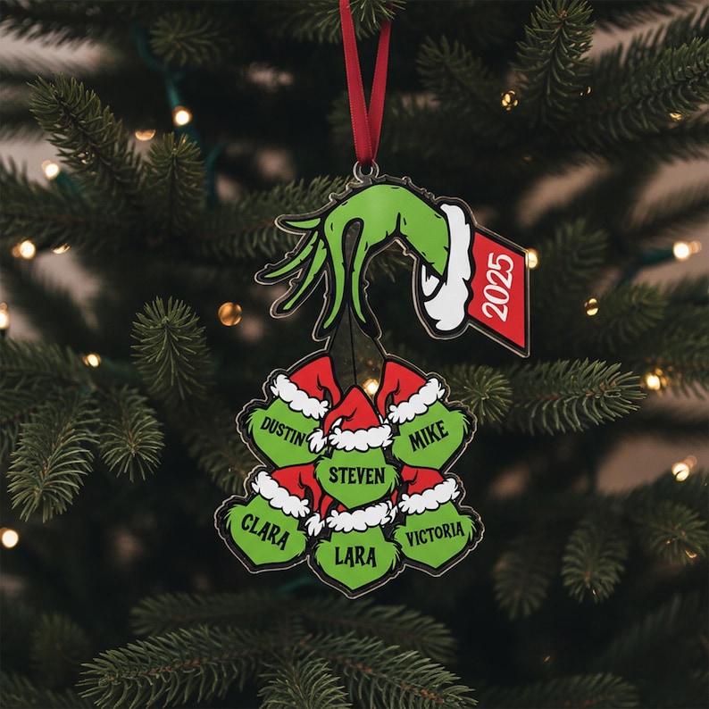 May include: A Christmas tree ornament featuring a green hand with a white cuff holding a cluster of red and white Santa hats. Each hat has a name on it. A red tag with the year 2025 hangs from the hand.