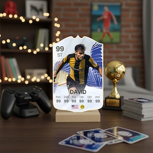Può includere: Una carta da calcio decorativa e illuminata con un giocatore di nome David. La carta mostra le statistiche del giocatore e il numero 99. La carta è montata su una base di legno. Altri oggetti a tema calcistico sono sullo sfondo.