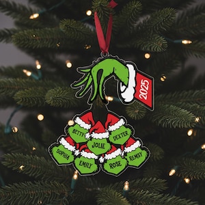 May include: A Christmas ornament featuring a green hand holding a cluster of red Santa hats, each with a name. The ornament has a red ribbon for hanging and a tag with the year 2025. The names include Betty, Dexter, Sophia, Emily, Jolie, Rose, and Remsy.
