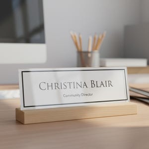 May include: A nameplate displaying "CHRISTINA BLAIR, Community Director" in black text on a white background, framed by a black border. It's held in a clear acrylic stand atop a light wooden base, suitable for an office desk.