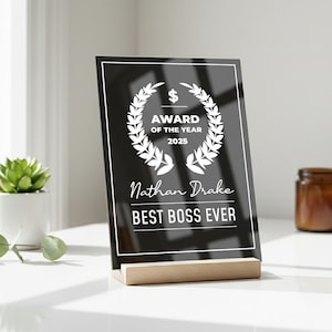 Könnte beinhalten: Eine schwarze Acryl-Auszeichnung mit weißem Text und Lorbeerkranz-Design. Der Text lautet "AWARD OF THE YEAR 2025", "Nathan Drake" und "BEST BOSS EVER". Die Auszeichnung wird auf einem Holzsockel präsentiert.