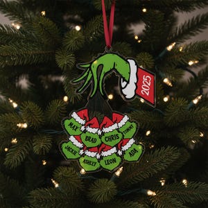 May include: A Christmas ornament featuring a green hand holding a cluster of green ornaments with names and red Santa hats. The ornament has a red ribbon for hanging and a tag that reads "2025".