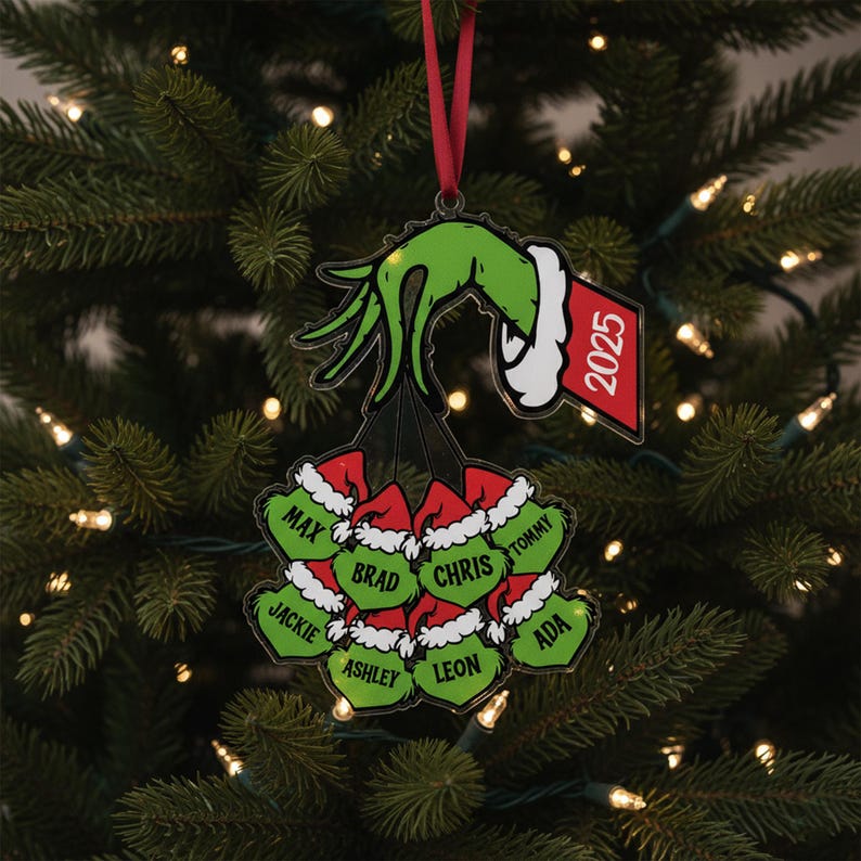 May include: A Christmas ornament featuring a green hand holding a cluster of green ornaments with names and red Santa hats. The ornament has a red ribbon for hanging and a tag that reads "2025".