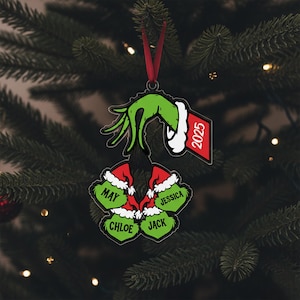 May include: A Christmas ornament featuring a green hand holding a wreath of four green ornaments with names: May, Jessica, Chloe, and Jack. The ornament includes a red tag with the year 2025.
