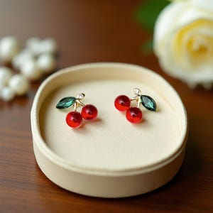 Puede incluir: Pendientes de cereza con cuentas de cereza rojas y brillantes, detalles de hojas verdes y pequeños detalles de cristal. Los pendientes se exhiben en una bandeja redonda para joyas de color crema. Una rosa blanca está en el fondo.