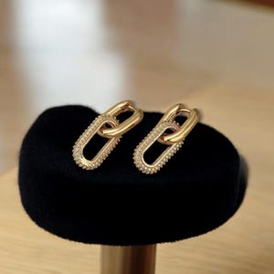 Pode incluir: Um par de brincos de argola dourados com um design de corrente ligada. Os brincos são texturizados com pequenos pontos em relevo.