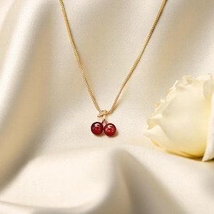 May include: A gold chain necklace with a cherry pendant. The pendant has two red, round cherry-shaped stones. The necklace is shown on cream-coloured satin fabric, with a white rose in the background.