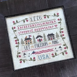 May include: A framed cross-stitch sampler with the text "Let Freedom Ring" and "1776" in a brown frame. The sampler features a red, white, and blue American flag, three houses, and the text "USA". The sampler is on a gray surface with red flowers.