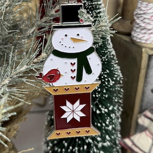 May include: A snowman Christmas ornament with a black top hat, green scarf, and a red bird. The ornament hangs from a red ribbon. The base is red with a white star design and gold trim. Festive holiday decor.