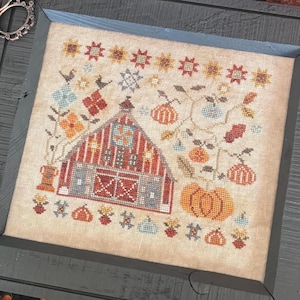 May include: A framed cross-stitch featuring a barn with pumpkins and fall foliage. Two knitted pumpkins, one white and one coral, sit beside the frame. A pair of decorative scissors rests near the frame. The background includes a fall-themed table runner.