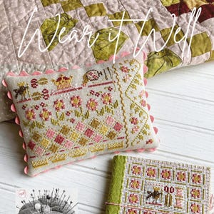 May include: A cross stitch project with the text "Wear it well" on a pink and green quilt. The project is a pillow with a floral design and a small book with a bird and floral design. The text "Pins & Needles" is on a black and white image of a pincushion.