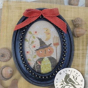 May include: An oval black frame containing a cross-stitch pattern of a pumpkin-headed figure wearing a witch hat, holding a broom, with a crescent moon and flowers. The frame is topped with a rustic orange ribbon bow and features text reading Hocus Pocus.