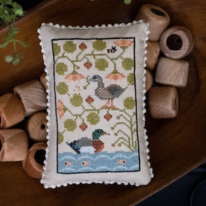 May include: A cross-stitch sampler featuring two ducks in a pond with a floral design. The sampler is titled "Quack Stack" and is an original design by Paulette Stewart. The sampler is displayed in a wooden bowl with spools of thread.
