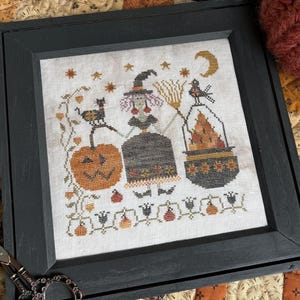 May include: A framed cross-stitch featuring Halloween-themed figures, including a witch, a black cat, and pumpkins. The artwork is in a black frame, with a pair of decorative scissors on the side. Two knitted pumpkins and a knitted white pumpkin are also in the image.